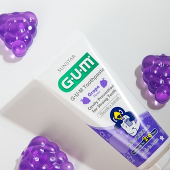 A tube of G.U.M toothpaste with grape flavor is positioned on a light surface. The packaging features purple and green accents, displaying information about cavity prevention and suitability for young children. Surrounding the toothpaste are plastic grape-shaped objects in a vibrant purple color.