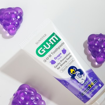 A tube of G.U.M toothpaste with grape flavor is positioned on a light surface. The packaging features purple and green accents, displaying information about cavity prevention and suitability for young children. Surrounding the toothpaste are plastic grape-shaped objects in a vibrant purple color.