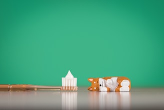 A small toy figure resembling a sleeping dog lies next to a toothbrush with toothpaste on its bristles. The background is a solid green color, and the objects are placed on a reflective surface.