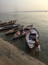 Several wooden boats are tied to a stone embankment by the riverside, with a few individuals attending to and managing the boats. The calm river stretches out to the horizon under a misty sky, creating a serene atmosphere. Steps lead down to the water, indicating a landing area or a ghat.