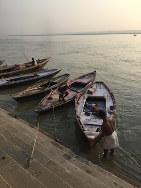 Several wooden boats are tied to a stone embankment by the riverside, with a few individuals attending to and managing the boats. The calm river stretches out to the horizon under a misty sky, creating a serene atmosphere. Steps lead down to the water, indicating a landing area or a ghat.