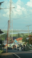 A rural scene with a tall communication tower, houses with colorful roofs, and several people riding motorcycles on a winding road. In the background, expansive fields stretch towards the horizon beneath a blue sky dotted with fluffy clouds.