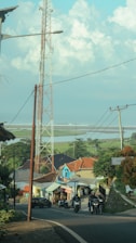 A rural scene with a tall communication tower, houses with colorful roofs, and several people riding motorcycles on a winding road. In the background, expansive fields stretch towards the horizon beneath a blue sky dotted with fluffy clouds.