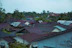 An aerial view of a residential area with numerous houses featuring rusty metal roofs, surrounded by lush green trees and palm trees visible in the background.