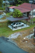 A silver sedan is parked under a simple metal carport with a brown roof beside a house. The background features a red-roofed house surrounded by greenery. The area around the driveway is a mix of grass, patches of gravel, and a paved road. A lush tree partially obscures the front of the house. The scene suggests a rural or suburban setting.