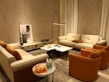 A cozy living room with modern furniture, featuring two beige sofas and two orange accent chairs arranged around a marble coffee table. A tall floor lamp provides lighting, while a piece of abstract art leans against the wall. Several decorative objects, including small sculptures and glass bottles on a side table, complete the setting.