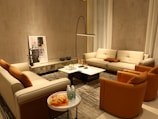 A cozy living room with modern furniture, featuring two beige sofas and two orange accent chairs arranged around a marble coffee table. A tall floor lamp provides lighting, while a piece of abstract art leans against the wall. Several decorative objects, including small sculptures and glass bottles on a side table, complete the setting.