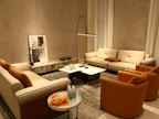 A cozy living room with modern furniture, featuring two beige sofas and two orange accent chairs arranged around a marble coffee table. A tall floor lamp provides lighting, while a piece of abstract art leans against the wall. Several decorative objects, including small sculptures and glass bottles on a side table, complete the setting.