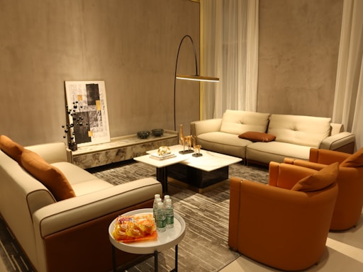 A cozy living room with modern furniture, featuring two beige sofas and two orange accent chairs arranged around a marble coffee table. A tall floor lamp provides lighting, while a piece of abstract art leans against the wall. Several decorative objects, including small sculptures and glass bottles on a side table, complete the setting.