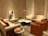 A cozy living room with modern furniture, featuring two beige sofas and two orange accent chairs arranged around a marble coffee table. A tall floor lamp provides lighting, while a piece of abstract art leans against the wall. Several decorative objects, including small sculptures and glass bottles on a side table, complete the setting.