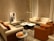A cozy living room with modern furniture, featuring two beige sofas and two orange accent chairs arranged around a marble coffee table. A tall floor lamp provides lighting, while a piece of abstract art leans against the wall. Several decorative objects, including small sculptures and glass bottles on a side table, complete the setting.