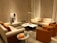 A cozy living room with modern furniture, featuring two beige sofas and two orange accent chairs arranged around a marble coffee table. A tall floor lamp provides lighting, while a piece of abstract art leans against the wall. Several decorative objects, including small sculptures and glass bottles on a side table, complete the setting.