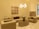 A stylish living room with beige and brown furniture. The room features a plush sofa set, an oval wooden coffee table adorned with a vase of white flowers, and a modern chandelier providing warm lighting. Wall decor includes a series of framed geometric art pieces.
