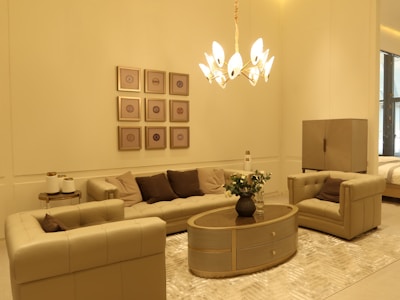 A stylish living room with beige and brown furniture. The room features a plush sofa set, an oval wooden coffee table adorned with a vase of white flowers, and a modern chandelier providing warm lighting. Wall decor includes a series of framed geometric art pieces.
