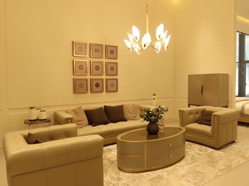 A stylish living room with beige and brown furniture. The room features a plush sofa set, an oval wooden coffee table adorned with a vase of white flowers, and a modern chandelier providing warm lighting. Wall decor includes a series of framed geometric art pieces.