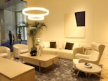 A modern, elegantly decorated living room with contemporary furniture. The space features a plush sofa with dark green cushions, complemented by a soft armchair and two cushioned chairs. A rectangular coffee table occupies the center, adorned with decorative items. Above, three circular pendant lights cast a warm glow. A modern abstract painting hangs on the wall, and a tall potted plant adds greenery. The room exudes a minimalist and sophisticated aesthetic.