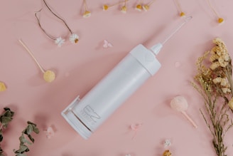 A white, cylindrical cleaning device with a long nozzle lies on a light pink surface. Surrounding it are various dried flowers and petals in shades of white, yellow, and green, arranged artistically.