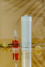 A small bottle with red liquid labeled 'RIINZ' is placed next to a cylindrical, white device labeled 'EZ Flosser' on a reflective surface, through which their mirrored images are visible. A decorative branch with yellow, spherical flowers and green leaves lies in the background, adding a natural element to the composition.