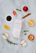 A flat lay composition featuring a variety of natural ingredients and a product box on a marble background. Visible items include sliced apples, orange halves, dry tea leaves on a small white dish, a small dropper bottle with a red liquid, cinnamon sticks, mint leaves, a sprig of rosemary, and a ripe red apple.