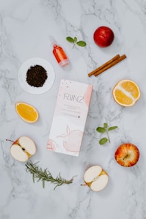 A flat lay composition featuring a variety of natural ingredients and a product box on a marble background. Visible items include sliced apples, orange halves, dry tea leaves on a small white dish, a small dropper bottle with a red liquid, cinnamon sticks, mint leaves, a sprig of rosemary, and a ripe red apple.