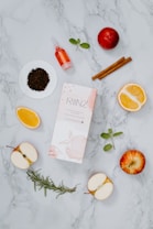 A flat lay composition featuring a variety of natural ingredients and a product box on a marble background. Visible items include sliced apples, orange halves, dry tea leaves on a small white dish, a small dropper bottle with a red liquid, cinnamon sticks, mint leaves, a sprig of rosemary, and a ripe red apple.