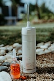 A portable water flosser and a small bottle of red liquid are placed outdoors on pebbly ground with some grass. The flosser is white with a nozzle on top and labeled EZ Flosser. The liquid bottle has a dropper lid and the word RINZ printed on it.