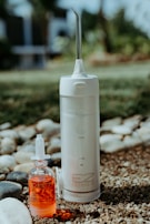 A portable water flosser and a small bottle of red liquid are placed outdoors on pebbly ground with some grass. The flosser is white with a nozzle on top and labeled EZ Flosser. The liquid bottle has a dropper lid and the word RINZ printed on it.