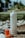 A portable water flosser and a small bottle of red liquid are placed outdoors on pebbly ground with some grass. The flosser is white with a nozzle on top and labeled EZ Flosser. The liquid bottle has a dropper lid and the word RINZ printed on it.