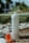 A portable water flosser and a small bottle of red liquid are placed outdoors on pebbly ground with some grass. The flosser is white with a nozzle on top and labeled EZ Flosser. The liquid bottle has a dropper lid and the word RINZ printed on it.