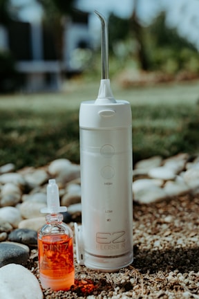 A portable water flosser and a small bottle of red liquid are placed outdoors on pebbly ground with some grass. The flosser is white with a nozzle on top and labeled EZ Flosser. The liquid bottle has a dropper lid and the word RINZ printed on it.