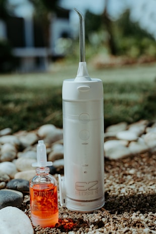 A portable water flosser and a small bottle of red liquid are placed outdoors on pebbly ground with some grass. The flosser is white with a nozzle on top and labeled EZ Flosser. The liquid bottle has a dropper lid and the word RINZ printed on it.