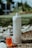 A portable water flosser and a small bottle of red liquid are placed outdoors on pebbly ground with some grass. The flosser is white with a nozzle on top and labeled EZ Flosser. The liquid bottle has a dropper lid and the word RINZ printed on it.