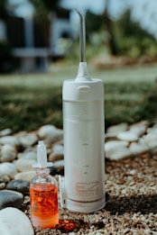 A portable water flosser and a small bottle of red liquid are placed outdoors on pebbly ground with some grass. The flosser is white with a nozzle on top and labeled EZ Flosser. The liquid bottle has a dropper lid and the word RINZ printed on it.