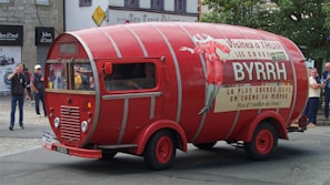 A vintage vehicle designed to look like a large red wine barrel is prominently displayed on a street. The vehicle features promotional signage for 'Byrrh,' with several people observing it in the background. The setting appears to be an urban street with stone buildings and shopfronts.