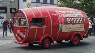 A vintage vehicle designed to look like a large red wine barrel is prominently displayed on a street. The vehicle features promotional signage for 'Byrrh,' with several people observing it in the background. The setting appears to be an urban street with stone buildings and shopfronts.