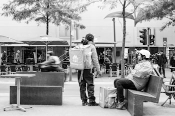 A grayscale street scene featuring two delivery workers with large square backpacks, branded 'Takeaway.com'. One stands while the other sits on a low bench, both dressed in outdoor gear. The background shows outdoor seating with umbrellas, a few pedestrians, and bicycles under leafless trees, suggesting an urban environment.