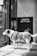 A dog with a white and brown coat stands patiently at the entrance of a store, its gaze directed through the glass door. The dog is wearing a harness and leash. Behind it, a sign with text in a foreign language is visible against the store front.