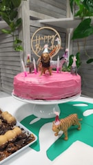 A pink frosted cake sits on a white cake stand, decorated with small animal figurines wearing party hats and surrounded by lit candles. A wooden 'Happy Birthday' topper is placed on the cake. In the foreground, a toy tiger with a party hat stands on a green paper cutout. To the side, there is a plate of desserts with chocolate and caramel toppings. The background features a wooden slat wall and some green foliage.