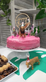 A pink frosted cake sits on a white cake stand, decorated with small animal figurines wearing party hats and surrounded by lit candles. A wooden 'Happy Birthday' topper is placed on the cake. In the foreground, a toy tiger with a party hat stands on a green paper cutout. To the side, there is a plate of desserts with chocolate and caramel toppings. The background features a wooden slat wall and some green foliage.