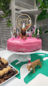 A pink frosted cake sits on a white cake stand, decorated with small animal figurines wearing party hats and surrounded by lit candles. A wooden 'Happy Birthday' topper is placed on the cake. In the foreground, a toy tiger with a party hat stands on a green paper cutout. To the side, there is a plate of desserts with chocolate and caramel toppings. The background features a wooden slat wall and some green foliage.