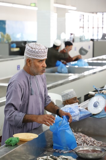 A man wearing traditional headgear stands at a fish market counter, packaging fresh fish into a blue plastic bag. Several other men in similar attire work at adjacent counters, some weighing fish or sorting through their stock. The environment is bright and functional with metallic counters and scales.