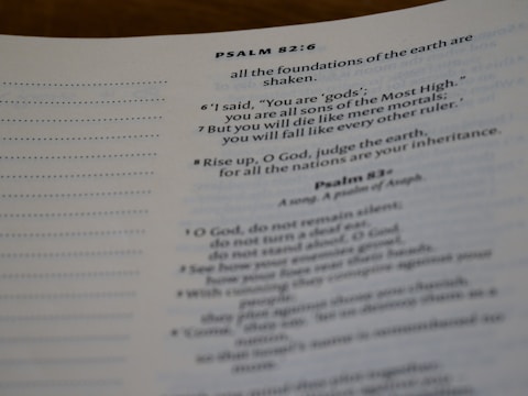Close-up view of a page from the Bible, focusing on Psalm 82:6-8. The text discusses themes of divinity and mortality. The page is slightly curled, and the paper appears off-white with printed text in a dark font.