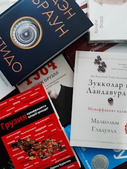 A collection of books arranged in a scattered manner with various covers visible, featuring texts in different languages. Some book covers are brightly colored, such as red and blue, while others are more neutral. Titles and designs vary, indicating a diverse range of genres or topics.