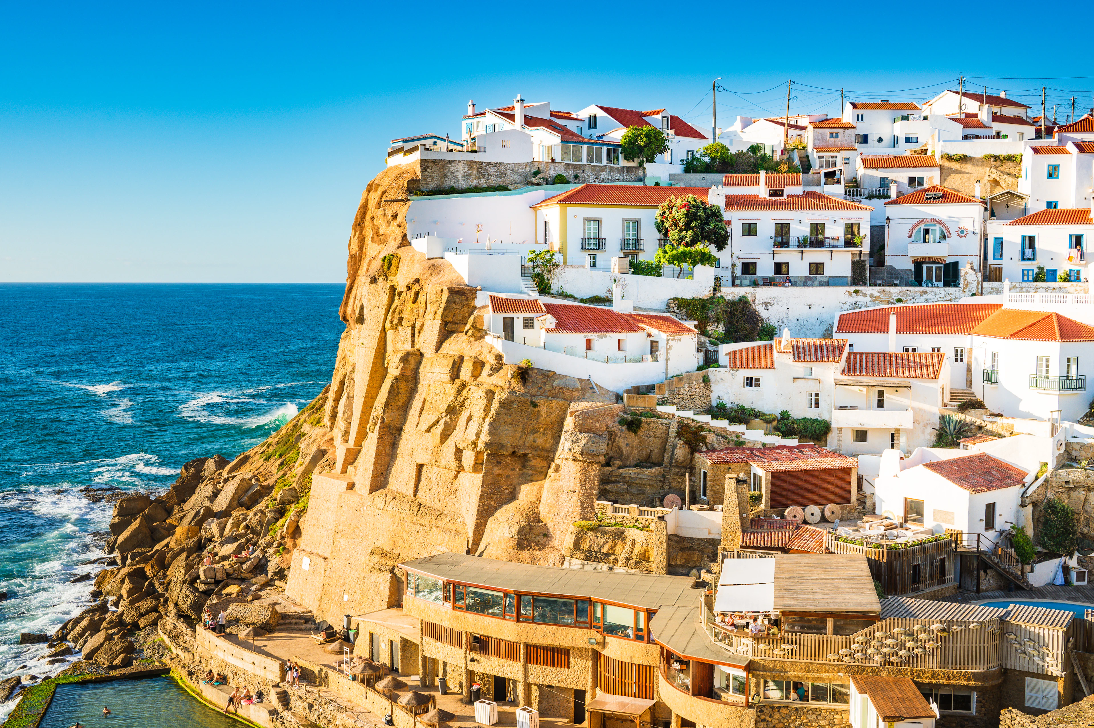 The white, red-roofed houses of Azenhas do Mar spilling down an ochre cliff above the Atlantic on the Colares coast, with the village's tidal sea pool cut into the rock at the foot of the headland.
