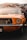 A vintage orange car with a classic design is prominently displayed, showcasing its front grille and one headlight. The background features a colorful, abstract mural painted on a wall.