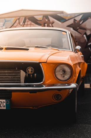 A vintage orange car with a classic design is prominently displayed, showcasing its front grille and one headlight. The background features a colorful, abstract mural painted on a wall.