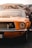 A vintage orange car with a classic design is prominently displayed, showcasing its front grille and one headlight. The background features a colorful, abstract mural painted on a wall.