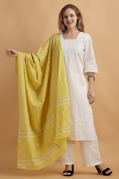 A person stands against a light brown backdrop wearing a white ethnic outfit with intricate patterns. They have a large, yellow scarf draped over the right shoulder, which features white designs. The person is smiling and wearing earrings and bracelets.