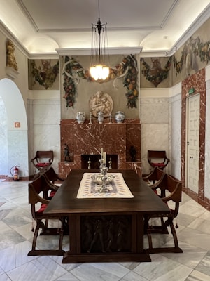 An elegant room features a large wooden dining table surrounded by ornate chairs. The table is adorned with a decorative table runner and a silver centerpiece with candlesticks. The walls are richly decorated with Renaissance-style frescoes and marble panels, and the ceiling boasts a classic chandelier providing warm illumination. Vases and sculptures accent the mantelpiece.