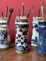 Several ornate ceramic jars, each decorated with intricate blue patterns on a white background, hold collections of paintbrushes. The jars are arranged on a wooden surface against a red wall.